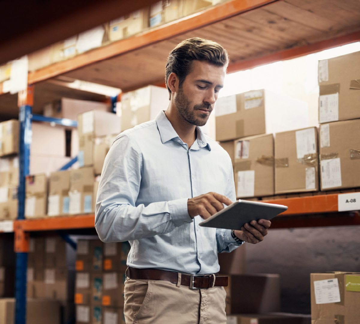 Man in warehouse using tablet – shelves of boxes in background.
